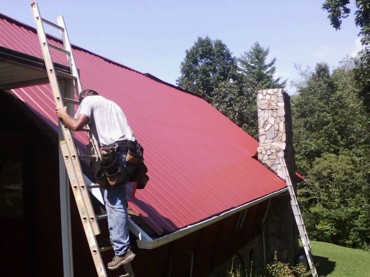 Roofer on a ladder beside a red metal roof and stone chimney — Soffit & Fascia Repair by Roofing Brothers in Wampum